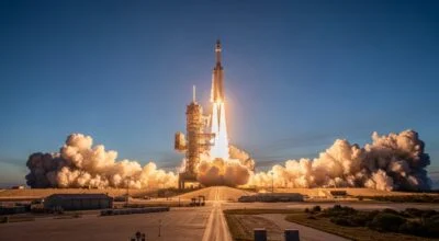 A rocket launches from a ground-based launch pad, ascending rapidly into a clear blue sky. Bright flames and exhaust burst from its engines, creating large billowing clouds of smoke on either side of the pad. The scene is captured from a distance, showing surrounding structures, flat terrain, and a long roadway leading toward the launch site, emphasizing the scale and power of the liftoff.