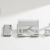 A clean, minimalist flat-lay of modern desk accessories arranged on a white surface. From left to right: a compact power adapter with a cable, a slim metallic power bank, a foldable aluminum laptop stand, a small pair of wireless earbuds in an open charging case, and a sleek white desk lamp. Soft lighting and neutral tones emphasize a tidy, contemporary workspace aesthetic.