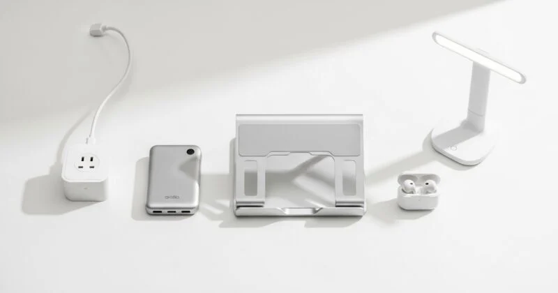 A clean, minimalist flat-lay of modern desk accessories arranged on a white surface. From left to right: a compact power adapter with a cable, a slim metallic power bank, a foldable aluminum laptop stand, a small pair of wireless earbuds in an open charging case, and a sleek white desk lamp. Soft lighting and neutral tones emphasize a tidy, contemporary workspace aesthetic.