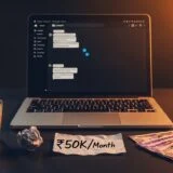 A dimly lit workspace scene featuring a laptop displaying a chat-style interface, suggesting online work or communication. In front of it, a handwritten note reads “₹50K/Month,” placed beside a small stack of Indian currency notes. A glass of hot coffee with visible steam sits to the left, along with a crumpled piece of paper. Warm light from one side contrasts with cooler tones on the other, creating a focused, late-night work atmosphere centered on earning or remote income.